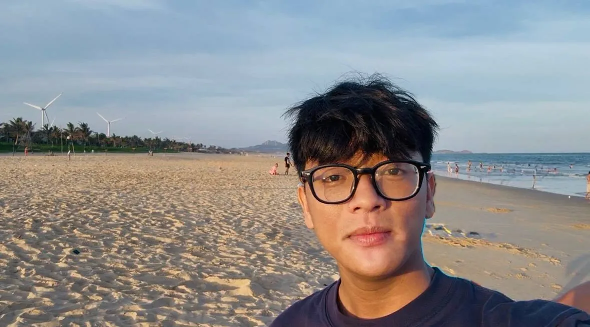 Đô Si U chụp selfie trên bãi biển lúc chiều muộn, phía xa là hàng cột điện gió